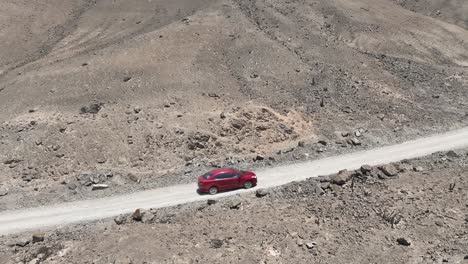 Toma-Panorámica-Aérea-De-Un-Vehículo-Rojo-Mientras-Viajaba-Por-Una-Carretera-Polvorienta-Y-Seca-En-Perú-A-Lo-Largo-De-Las-Colinas-Y-Piedras-En-El-Costado-Cerca-De-La-Ciudad-Santa-De-Caral-En-Un-Día-Soleado