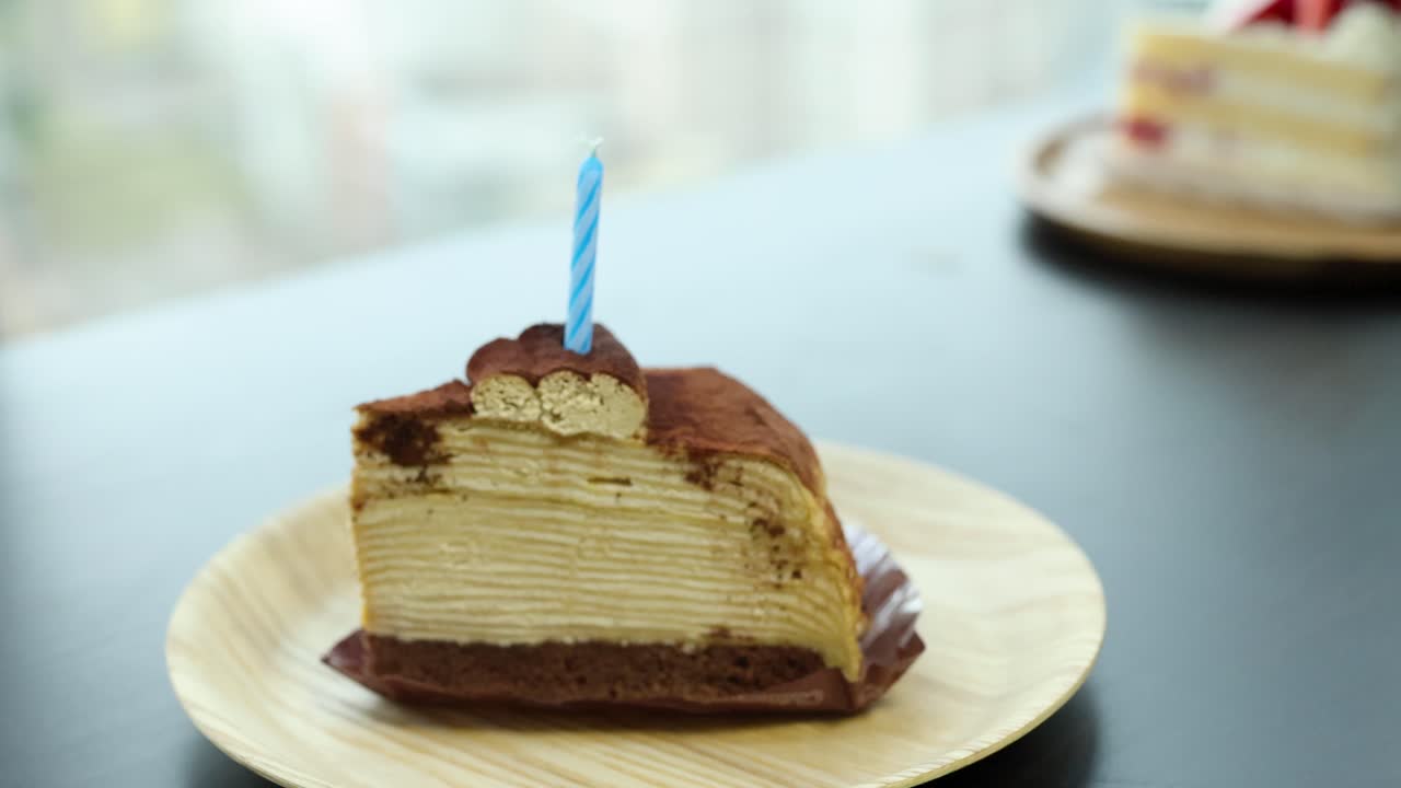 Hand inserts blue birthday candle into tiramisu crepe cake slice in soft natural daylight