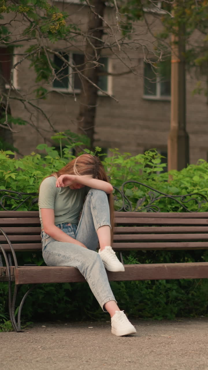 야외 공원에서 나무 벤치에 앉아있는 젊은 여성, 울창한 초록색과 나무로 둘러싸인 반사적이고 우울한 자세로 팔에 머리를 고 있습니다.