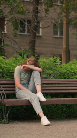 mujer joven sentada en un banco de madera en un parque al aire libre, descansando la cabeza en su brazo en una postura reflexiva y sombría, rodeada de exuberante vegetación y árboles