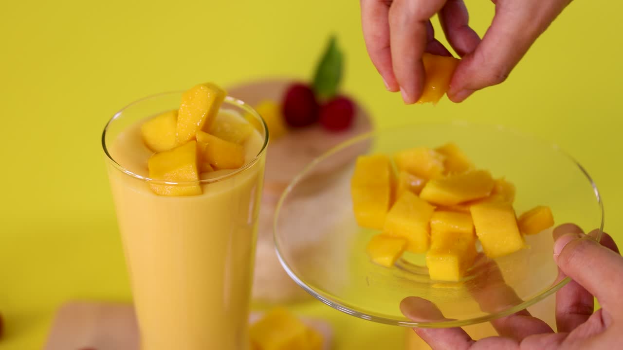 Hand places mango cubes on smoothie in glass, bright yellow background, natural lighting, static camera