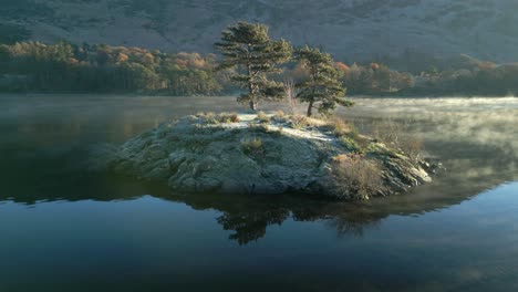 small rocky island with two trees on calm lake and mist curling around the island at sunrise in autumn
