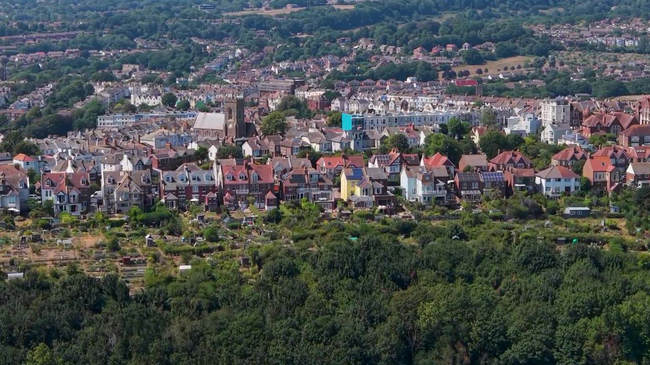 Approccio aereo di Belmont verso la chiesa di Emmanuel a Hastings, con case sulla collina circostanti, che mostra tetti residenziali densi, bordo boschivo frondoso e profondità del paesaggio urbano alla luce del giorno