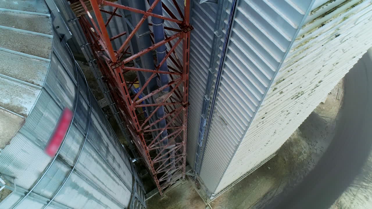 Metal brown tower with tube inside attached to a silo tank and warehouse. Descending footage along the granary premises. Top view.