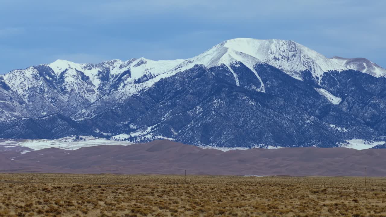 High-angle drone shot capturing the intersection of desert dunes and alpine peaks. The snow-covered slopes of Mount Herard rise sharply above the expansive Great Sand Dunes in Southern Colorado