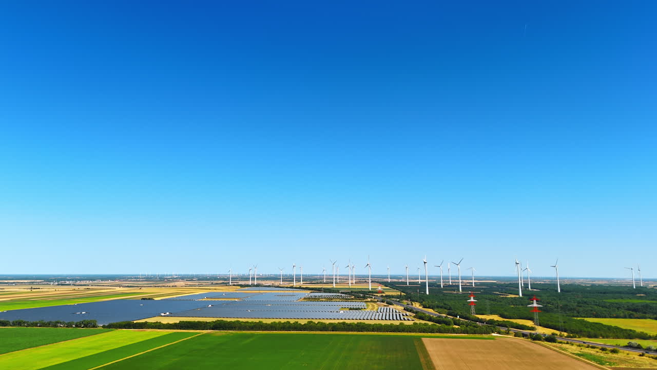 Approaching the modern sites of solar fields installed in the countryside. Wind farms work at backdrop. Aerial view