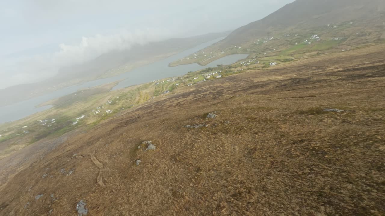 avión no tripulado volando sobre una cresta a través de nubes bajas hacia un pequeño pueblo junto al océano atlántico, irlanda