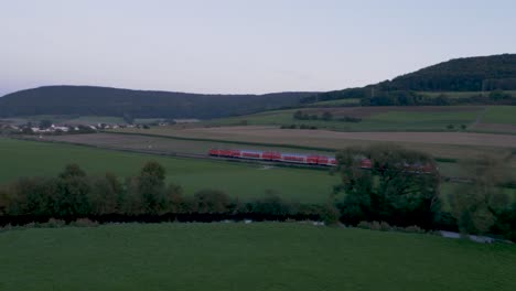 Drone-shot-at-dusk-reveals-a-red-train-with-several-passenger-cars,-moving-from-right-to-left-across-a-single-track-in-a-green-field,-a-quaint-village-with-white-houses,-under-a-pale-blue-sky