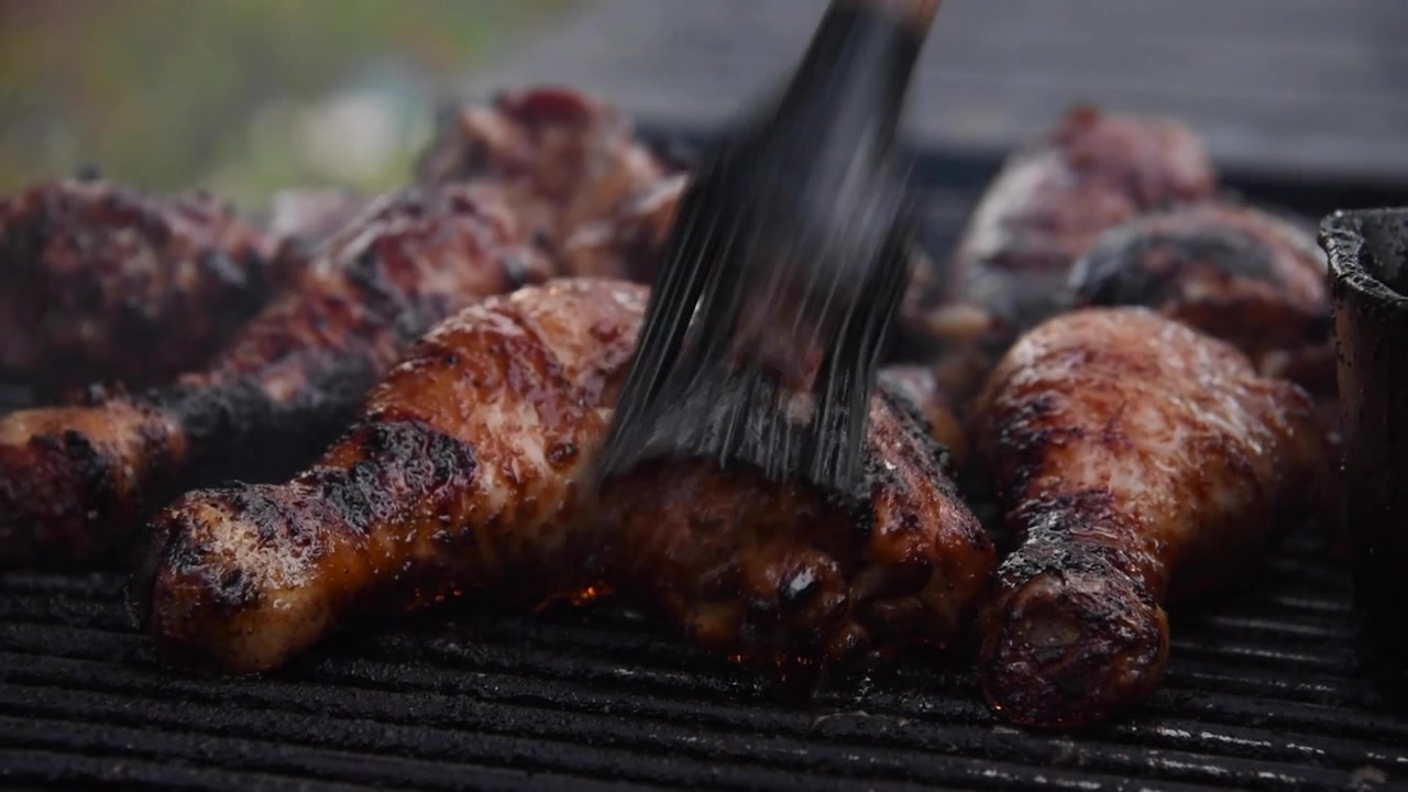 Spennellatura culinaria di cosce di pollo marinate con salsa barbecue. Il pollame sfrigola e fuma sulla griglia calda durante la cottura.