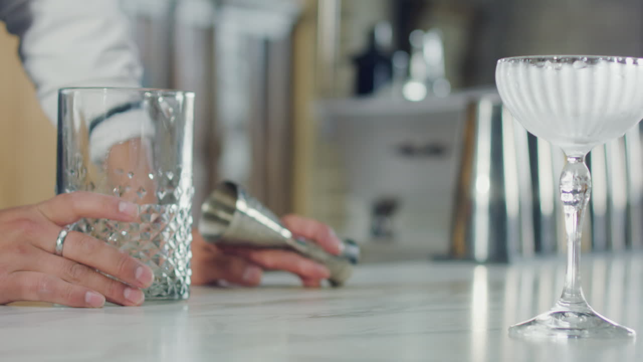 Close-up of a bartender arranging a glass next to a goblet, with a jigger out of focus in the background