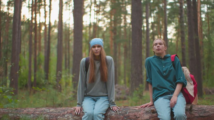 уставшие двоюродные братья с рюкзаками сидят на упавшем дереве в пышном лесу, один двоюродный брат, одетый в синюю бандану, счастливо поднимает ногу, кажусь расслабленным и радостным, в то время как двоюродной брат в зеленой рубашке опирается на руку на дерево