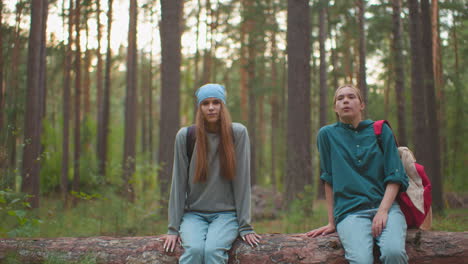 tired cousins with backpacks sit on fallen tree in lush forest, one cousin, wearing blue bandana, lifts leg happily, appearing relaxed and joyful, while cousin in green shirt rests hand on tree