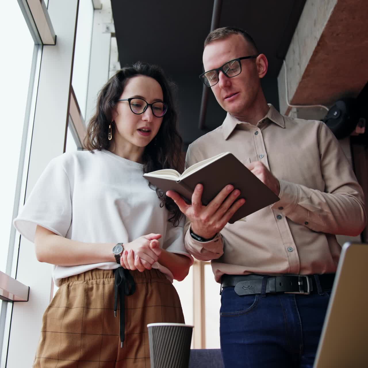People working together stand in the office. Teammates look through the paper notebook, work schedule or plans, discussing and revising. Low angle view