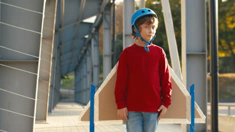 portrait of a cute little boy in helmet and red sweater looking at the camera while standing on a bridge on a sunny day 1