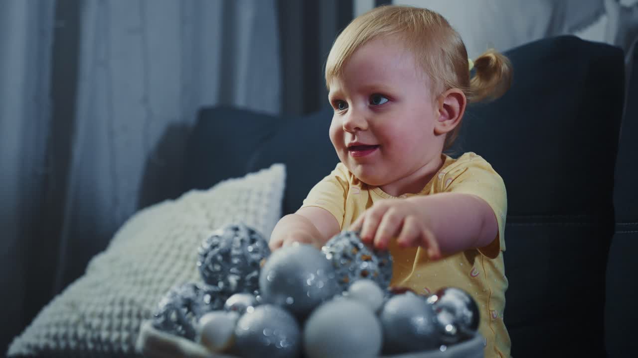 Adorable baby girl sitting on cozy sofa, receiving silver and blue christmas balls from caring mother's hand, exploring festive decorations with innocent curiosity and joyful smile