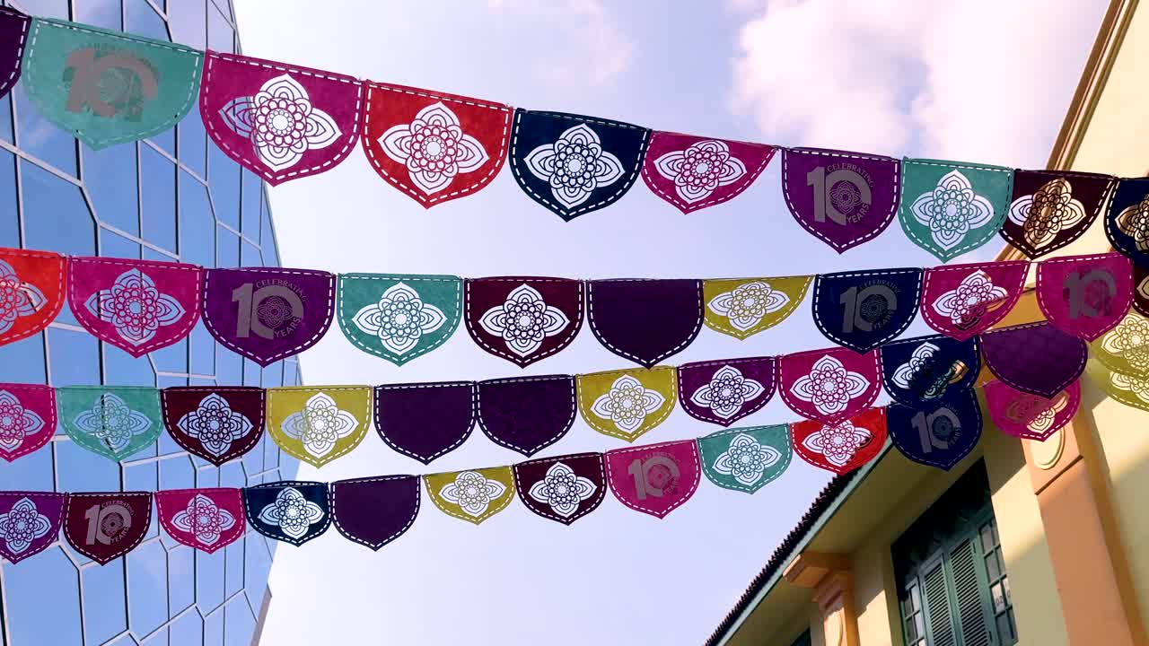 Rows of vibrant decorative flags flutter overhead between modern glass buildings and traditional architecture, captured with smooth upward camera movement in natural daylight