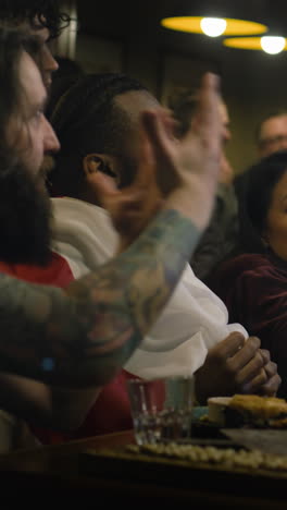 vertical shot of fans or friends watch football match broadcast and discuss game sitting in sports bar or pub at night. multi ethnic people support soccer team playing in world league championship.