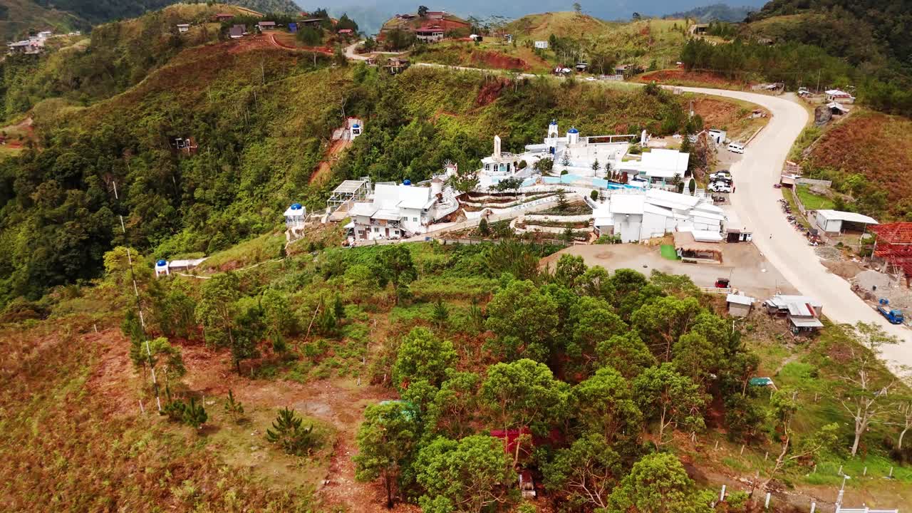 Aerial shot of whitewashed buildings in Greek style atop scenic highlands — surrounded by green forest, winding roads, and mountain views. Ideal for travel or real estate use
