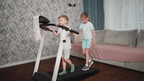 boy walks casually on treadmill while girl behind focuses on keeping balance during light indoor workout session in cozy room with pink covered couch, patterned wallpaper
