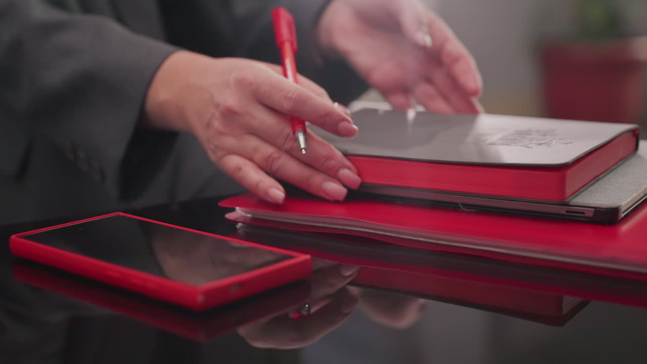 Nærbillede af forretningskvindes hånd, der placerer en rød smartphone oven på røde notesbøger og papirer på et blankt skrivebord. Organiseret virksomhedsarbejdsplads, der fremviser produktivitetsværktøjer og fokus på arbejde.