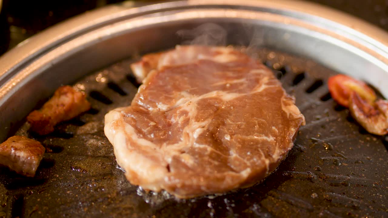 Raw pork steak sizzles on hot grill, surrounded by vegetables, under warm restaurant lighting
