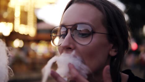 Beautiful-girl-taking-cotton-candy-in-and-amusement-park-near-the-blurred-carousel.-Wearing-trendy-glasses,-happy,-smiling.-Close