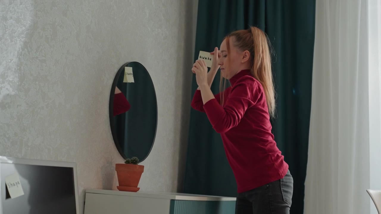 Giovane donna con un top rosso attacca post-it su uno specchio ovale. La donna tiene in mano diversi post-it gialli. La scena include pareti chiare e strutturate e tende verde scuro.