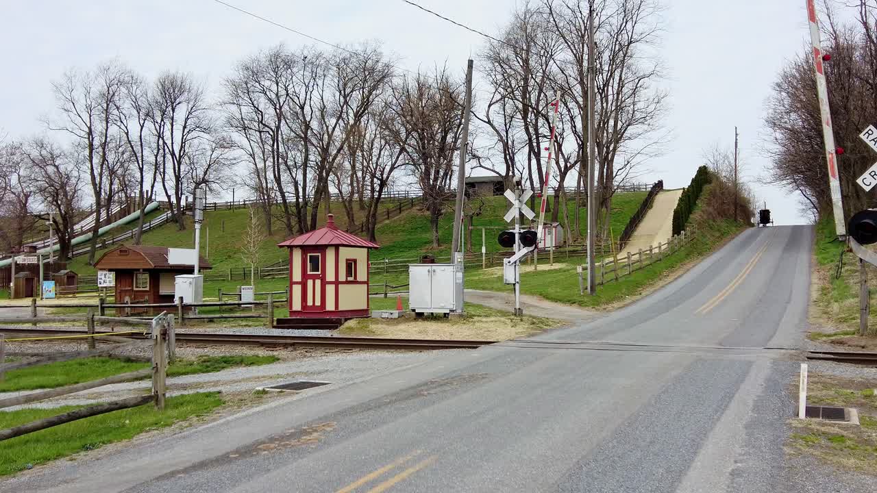 A horse-drawn carriage travels toward a railway crossing in the countryside. Surrounding structures consist of small buildings and trees, indicating a peaceful rural area.