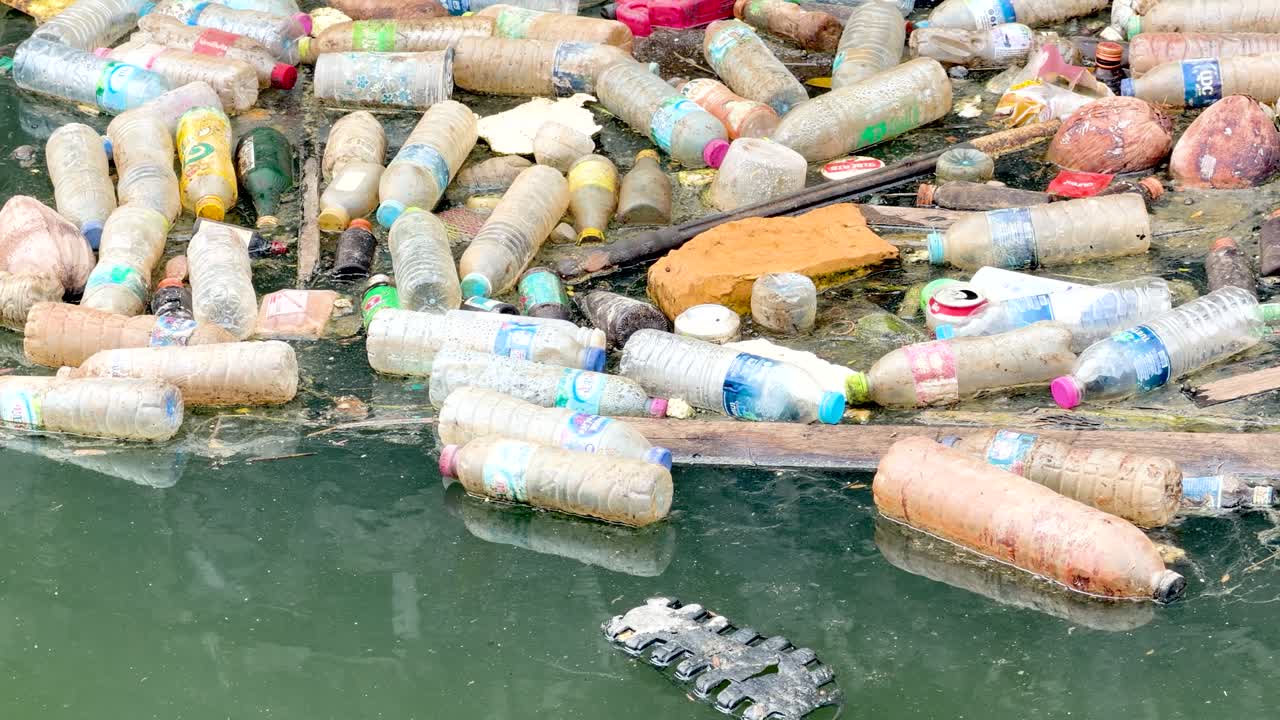 Inquadratura statica dall'alto di rifiuti di plastica e detriti che galleggiano sull'acqua verde e inquinata di un canale.