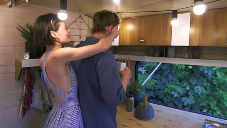 pareja joven positiva que vive o viaja en una furgoneta - la esposa viene y cierra los ojos de su marido por detrás. el hombre está preparando té en la cocina, dando caja de té para olerlo. acogedora, vida alegre juntos
