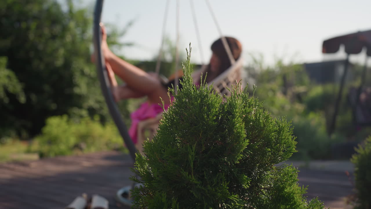 Tranquil Scene With Woman Reclining In Hanging Chair Amidst Nature, Laidback Woman Enjoys Quiet Moment Sitting In Shaded Outdoor Chair Surrounded By Lush Foliage And Gentle Sunshine