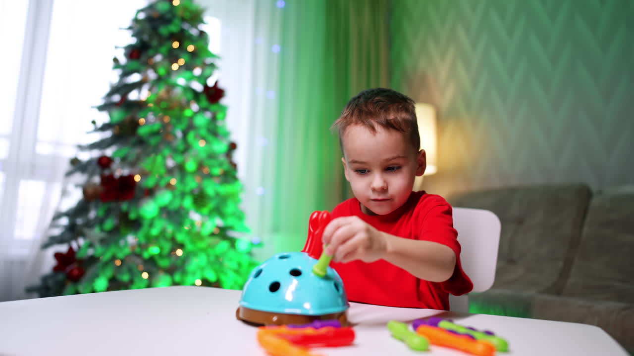 Lovely kid in red t-shirt plays with a toy sitting at desk. Cute child focused on a game at home. Christmas tree at backdrop.