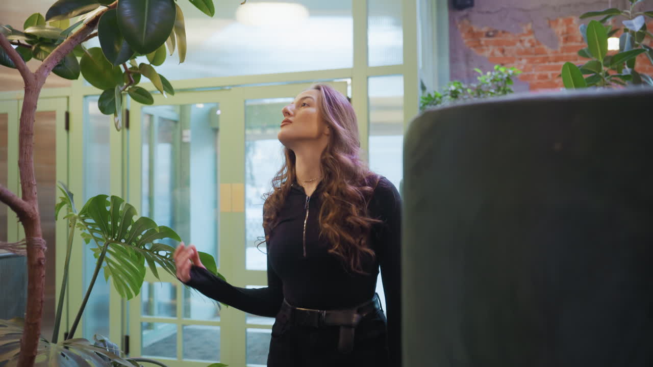 Stylish woman dressed in black walks confidently into cozy interior filled with lush indoor plants, gently brushing leaves as soft outdoor light reveals faint movement of vehicles through glass doors