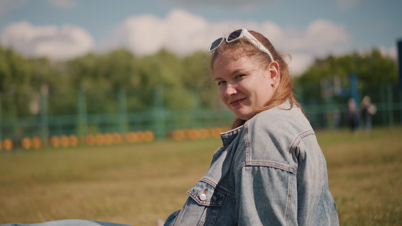 Nærbillede af en hvid kvinde, der smiler i denim, med en udendørs parkbaggrund med træer og solskin. Solbriller på hovedet og hårbånd, synlige fregner, et ægte engagerende udtryk og en glad, selvsikker stemning.