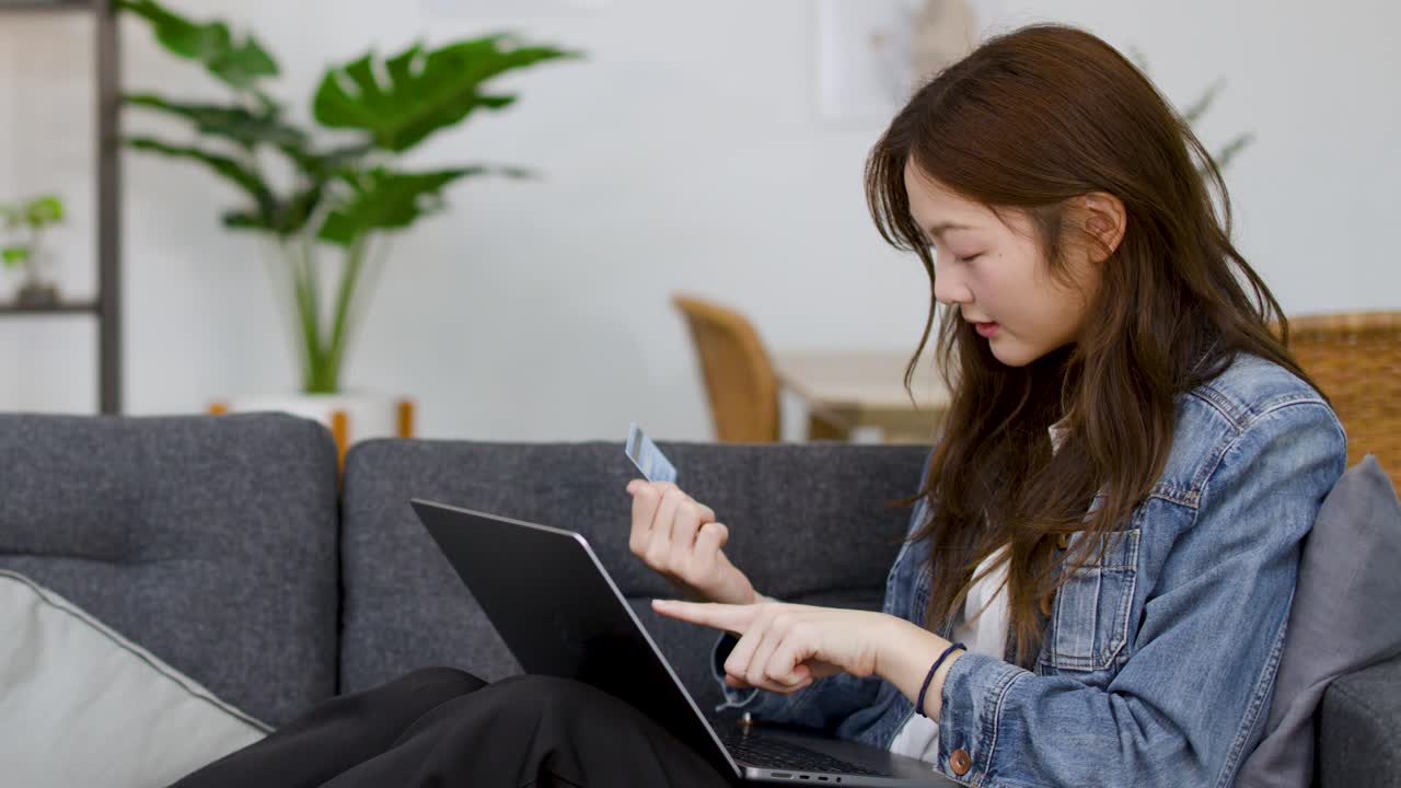 一位年轻女性坐在沙发上，使用笔记本电脑和信用卡购物。