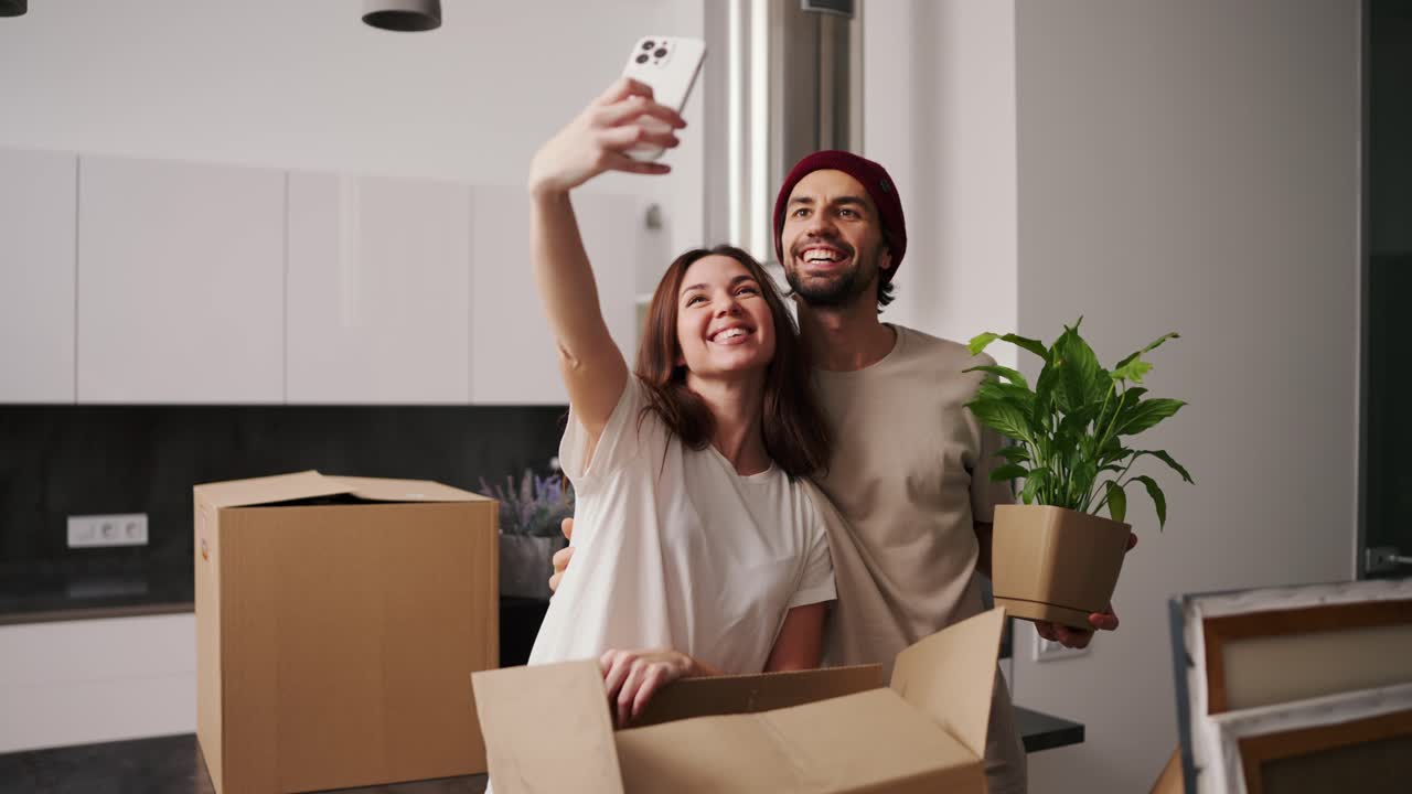 una chica morena feliz con una camiseta blanca toma una selfie usando un teléfono inteligente blanco con su novio con paja en una camisetta beige y plantas de la casa durante su mudanza entre un gran número de cajas en un apartamento moderno