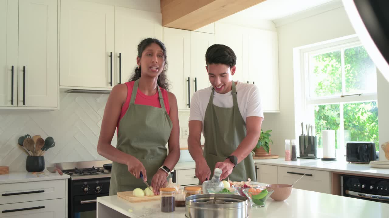 Casal afro-americano picando cebolas, ralando em uma panela, falando para um anel de luz na ilha da cozinha