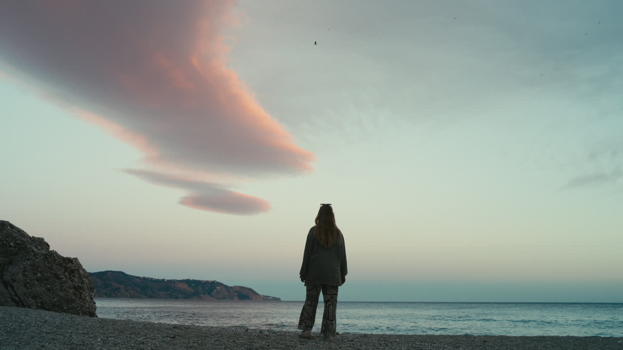 Vue de dos d'une femme immobile sur une plage de galets au crépuscule, contemplant la mer calme sous des nuages roses spectaculaires dans un large cadre cinématographique