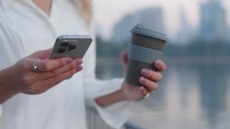 close-up of woman's hands holding a smartphone and a reusable coffee cup in an urban setting