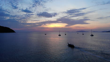 un barco de cuento tailandés navegando hacia el atardecer con otros barcos anclados