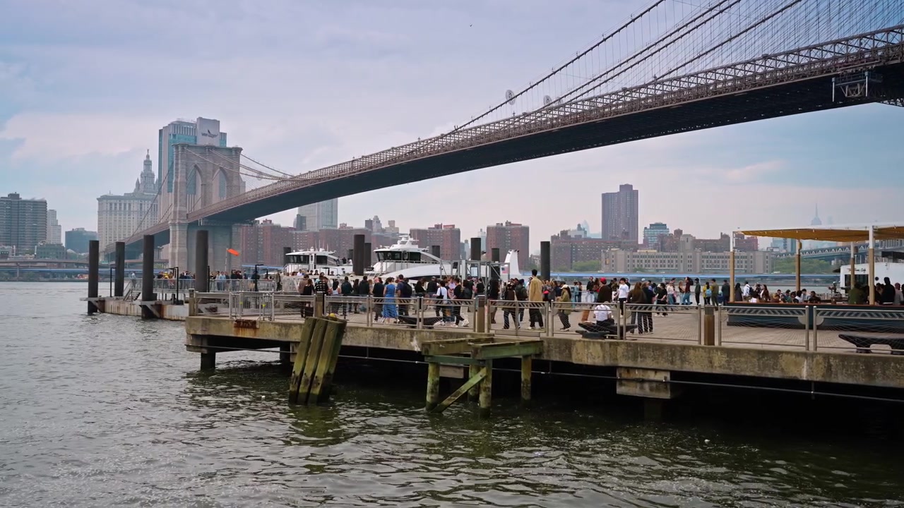 New York, USA, 1 maggio 2026: Folla sul molo lungomare sotto il Ponte di Brooklyn. Le persone si radunano su un ampio molo di legno con aree salotto sotto una massiccia struttura in acciaio del ponte.
