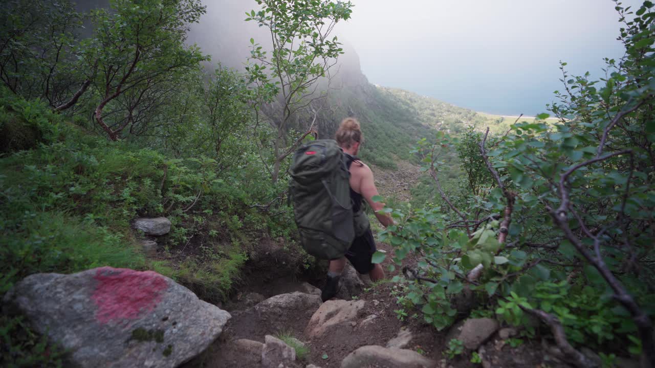 un excursionista masculino con una mochila de camping que desciende de un estrecho sendero rocoso en un día de niebla en el monte donnamannen, nordland, noruega