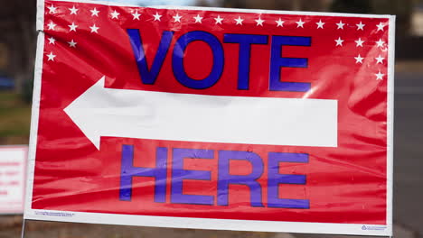 Vote-Here-Sign-Pointing-Left-with-People-Driving-Cars-in-Background,-Close-Up