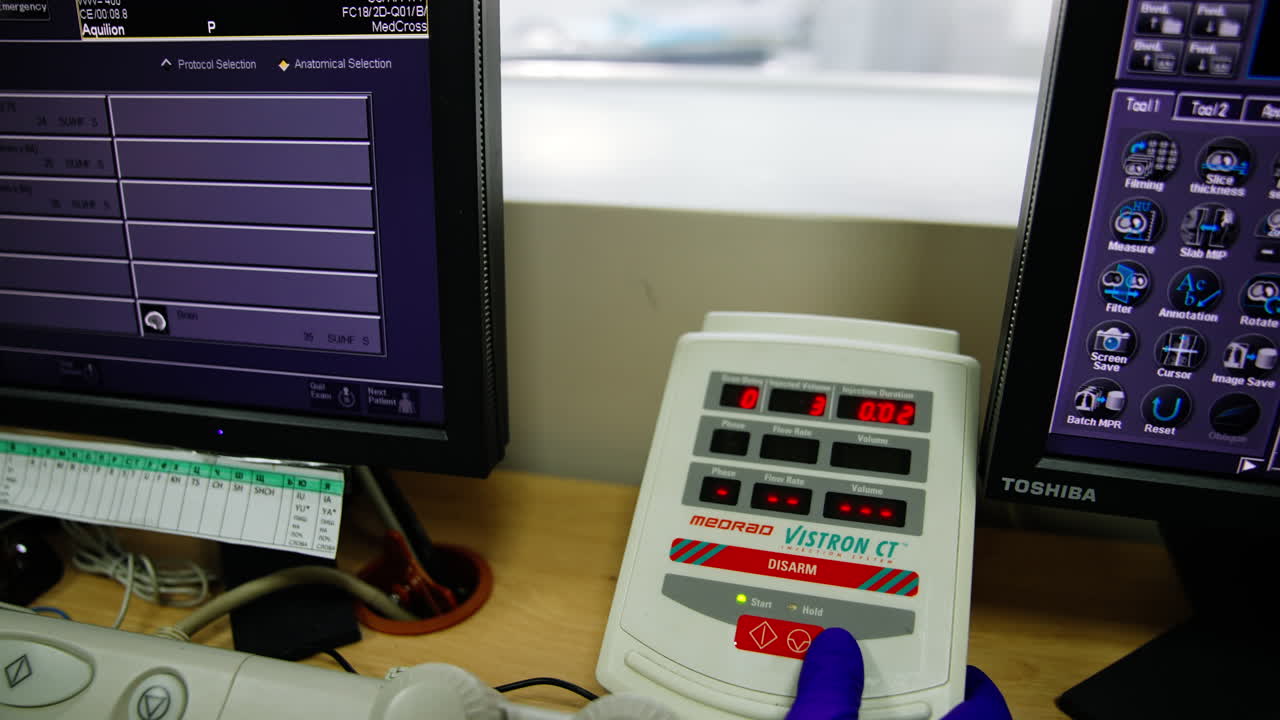 Lab technician's gloved hand presses buttons on the equipment panel near two working computer screens. Modern MRI machine at backdrop.
