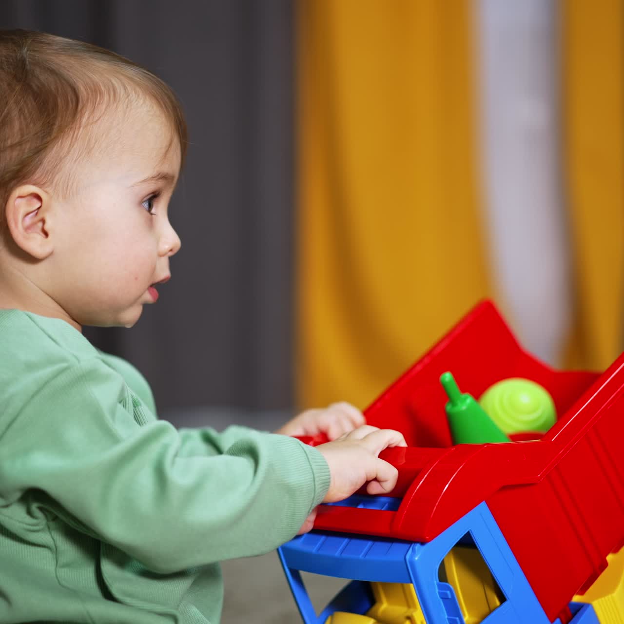 Little baby boy in light green shirt holding a big colorful lorry. Brown-haired kid playing with a toy in the room and looking around. Blurred backdrop