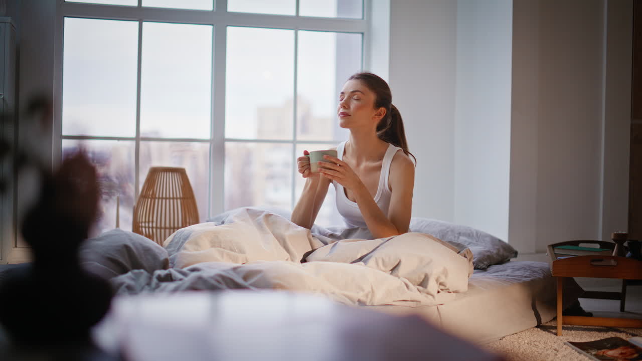 Ragazza appena sveglia che beve caffè in camera da letto, sentendosi calma a casa, al mattino, da sola.