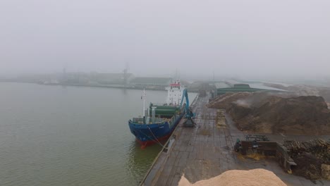 aerial establishing view of wood terminal crane loading timber into the cargo ship, port of liepaja , lumber log export, overcast day with fog and mist, wide drone shot moving forward slow