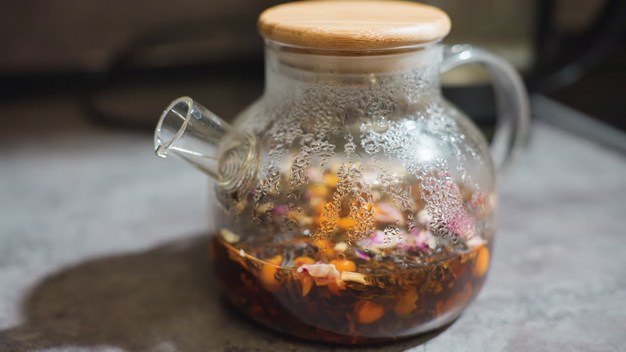 Steeping Herbal Tea Inside Glass Container, Cozy Scene Of Herbal Infusion Resting In Glass Teapot, Glass Teapot With Lid Shows Herbal Tea Steeping With Condensation And Scattered Petals
