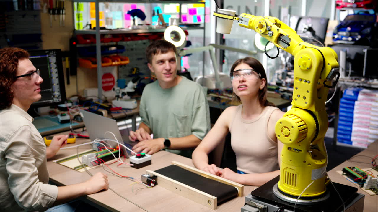 Young happy engineers programming an yellow robotic arm in the workshop to grab cardboard water glass, computer programming training for coffee preparation, celebrating success
