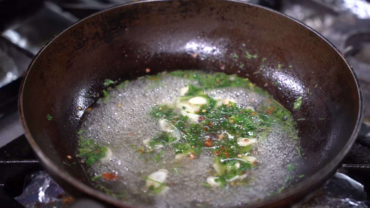 Un primo piano di fette d'aglio ed erbe fresche che sfrigolano in olio caldo in una padella rustica. La miscela crea una base aromatica per un piatto saporito in un ambiente di cucina professionale